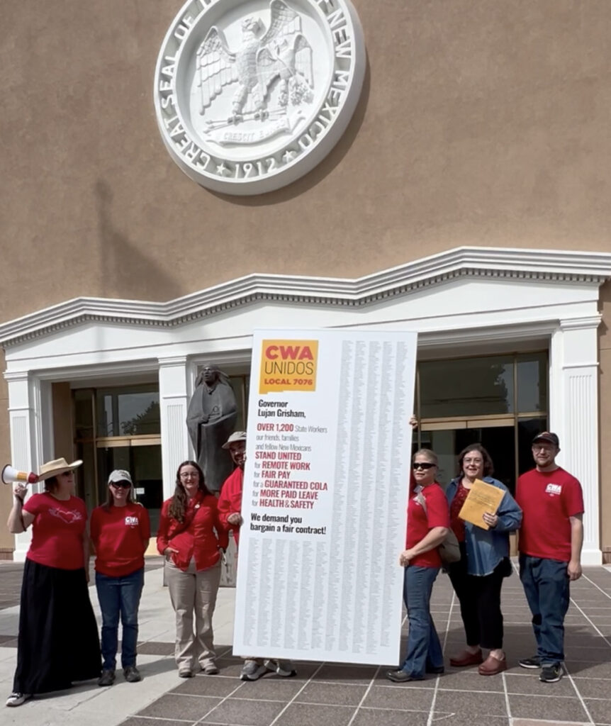 Members of the CWA Local 7076 State of New Mexico contract bargaining team, wearing red and holding a bullhorn, present a large sign in front of the Roundhouse. Over 1,200 state workers, our friends, families and fellow New Mexicans signed our petition demanding fair contract bargaining for remote work, fair pay, a guaranteed COLA, more paid leave, and health and safety.